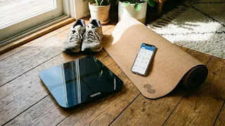 RENPHO smart body fat scale on a wooden floor with yoga equipment, displaying the Renpho app interface for tracking BMI and body fat percentage.