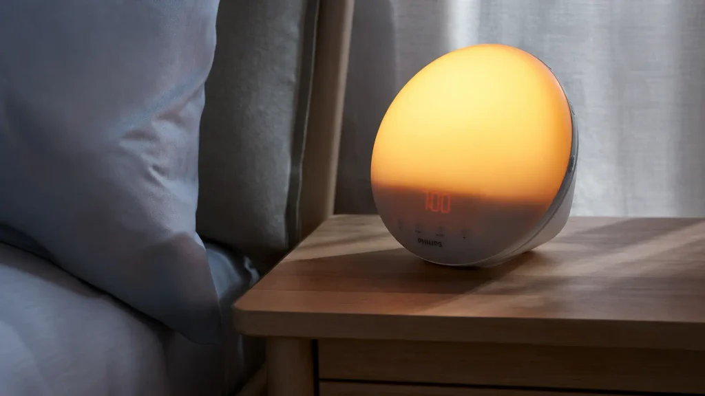 The Philips SmartSleep sunrise alarm clock for daylight saving emitting a bright, warm yellow glow on a bedroom nightstand during a dark morning.