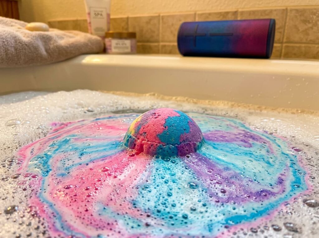 An Ultimate Ears Boom 3 waterproof Bluetooth speaker rests safely on a tub ledge next to a fizzing LifeAround2Angels bath bomb, creating the perfect audio and aromatherapy ecosystem for a spa night.
