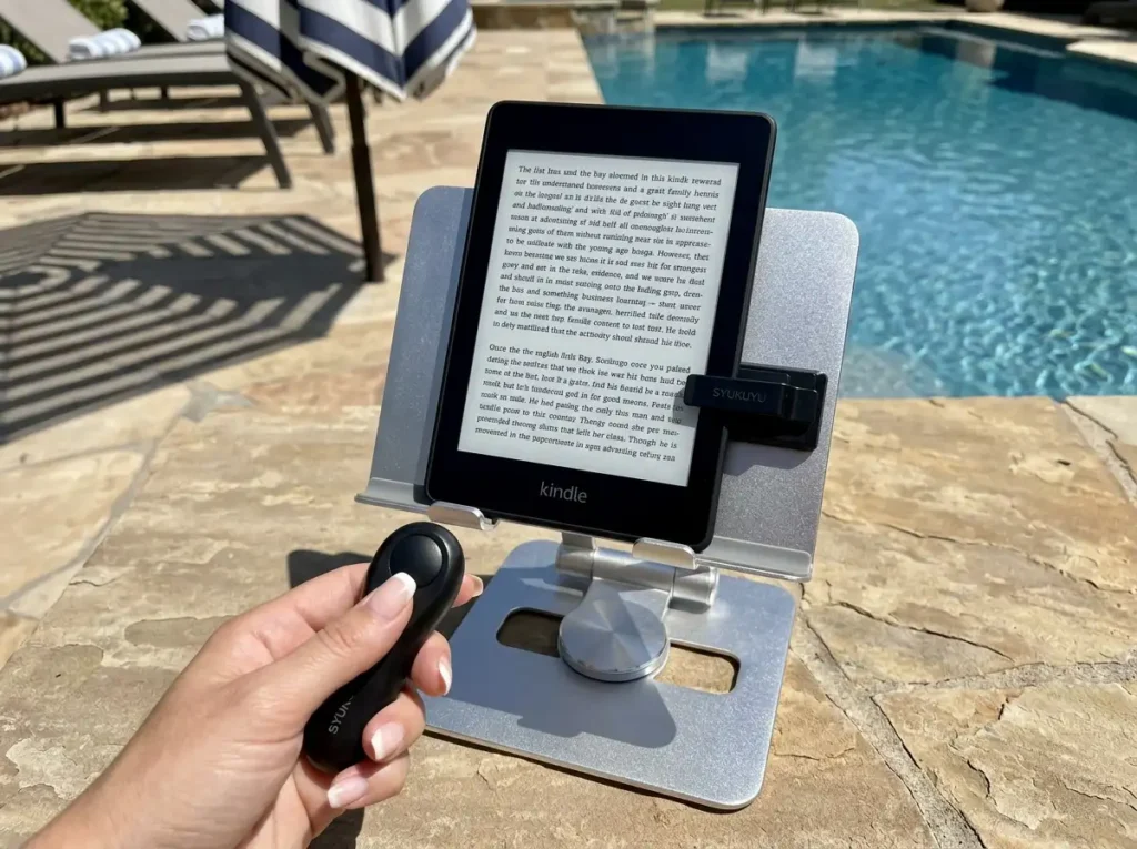 A SYUKUYU RF Remote Control Page Turner clipped to a Kindle Paperwhite on a pool stand, with a hand holding the small remote clicker.
