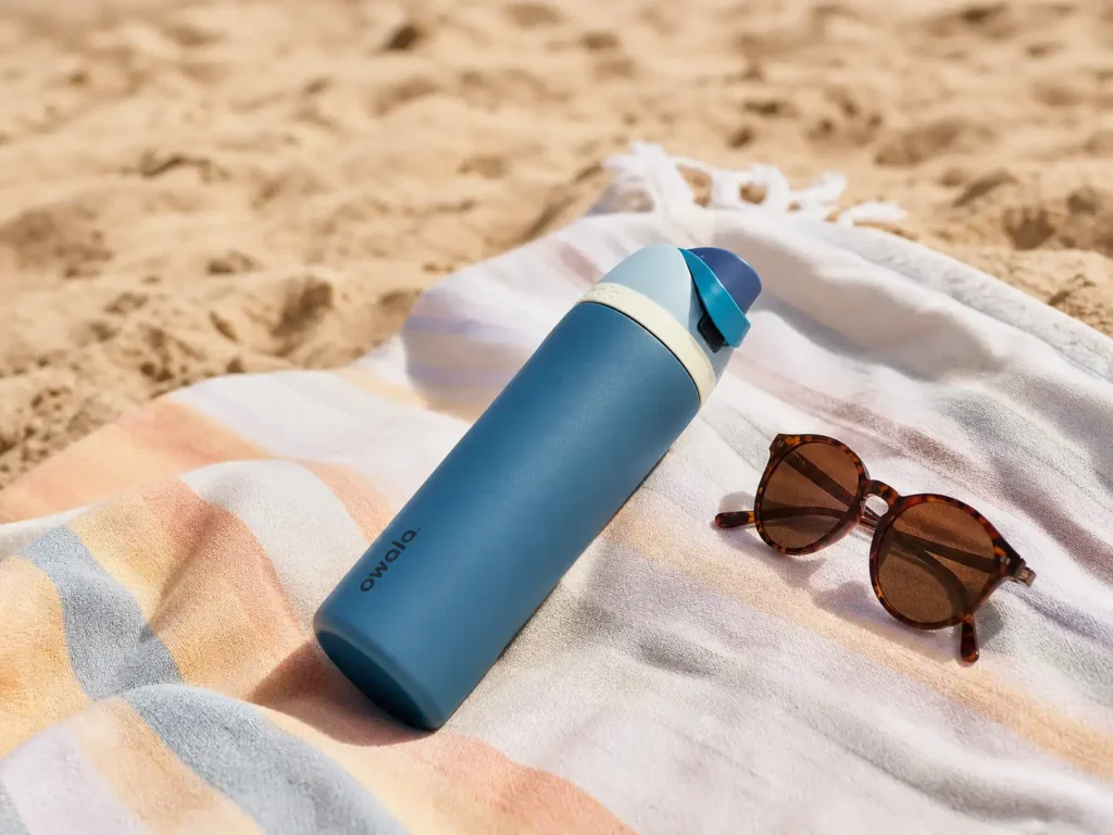 An Owala FreeSip Insulated Stainless Steel Water Bottle resting on a beach towel in the bright sun
