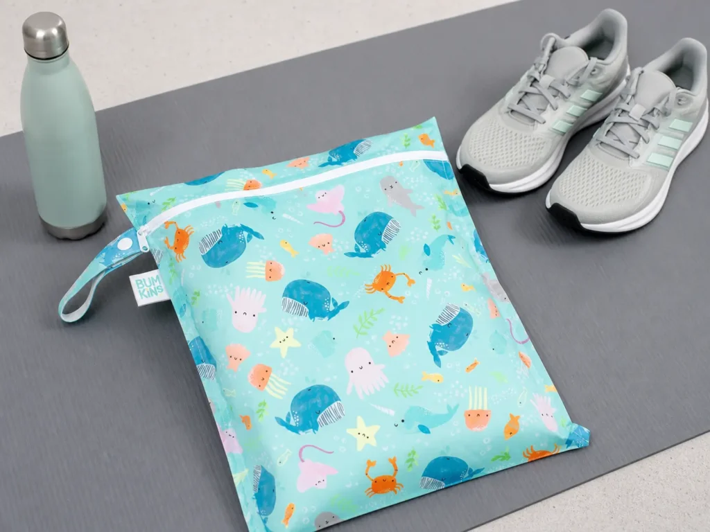 A Bumkins waterproof wet bag with tropical pattern lying next to a water bottle and gym shoes on a fitness mat.