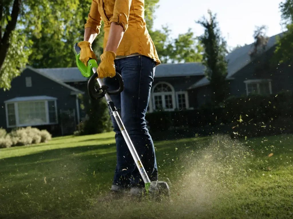 A homeowner neatly trimming overhanging lawn grass near a garden mulch bed with a green and black Greenworks 40V string trimmer