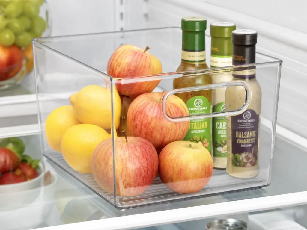 mDesign clear plastic storage bin with handles filled with fresh produce and condiments on a refrigerator shelf.