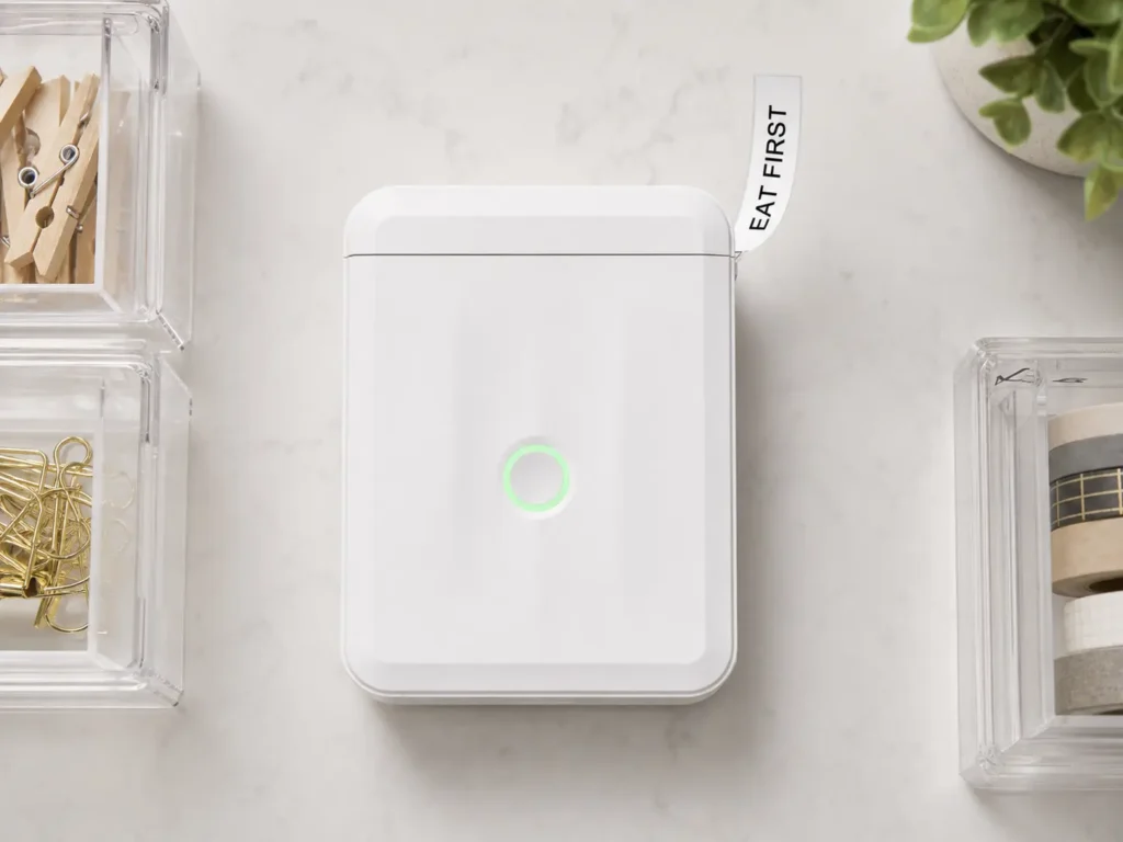 A compact white NIIMBOT D110 thermal label maker printing a label beside clear kitchen storage bins.