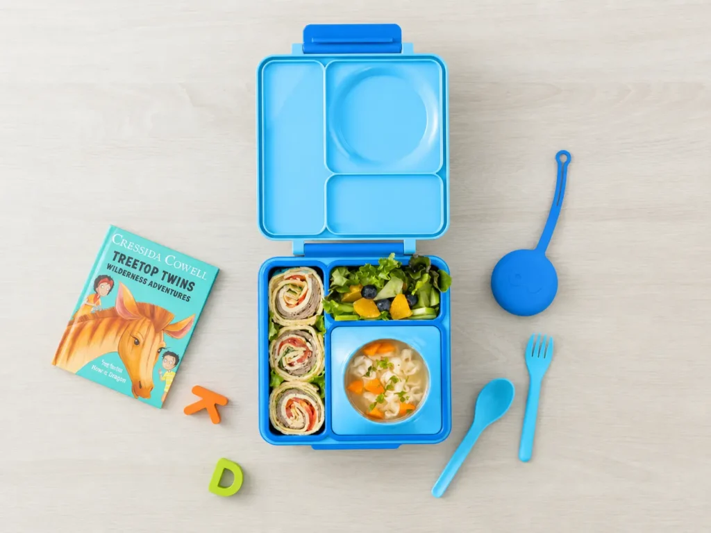 OmieBox insulated bento lunch box with its integrated stainless steel food jar open to show steaming warm chicken noodle soup next to sides of grapes and cucumber slices