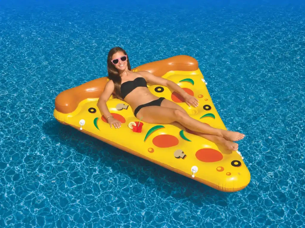 An inflatable Swimline pizza slice pool float with realistic pepperoni and veggie prints drifting in a clear backyard pool.